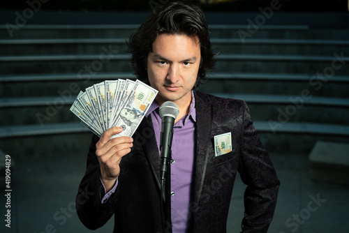 Male Asian Stand Up Comedian with Microphone and Fanned $100 Dollar Bills in Suit in Empty Auditorium Funny Look Face How to Making Money MLM Rich