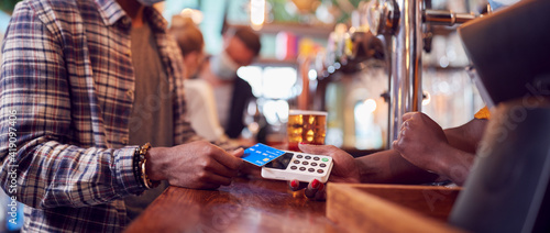 Male Customer Wearing Mask In Bar Making Contactless Payment For Drinks During Health Pandemic photo