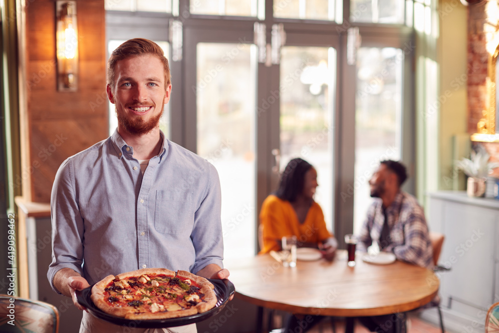 Portrait Of Waiter In Restaurant Serving Pizza To Couple At Table