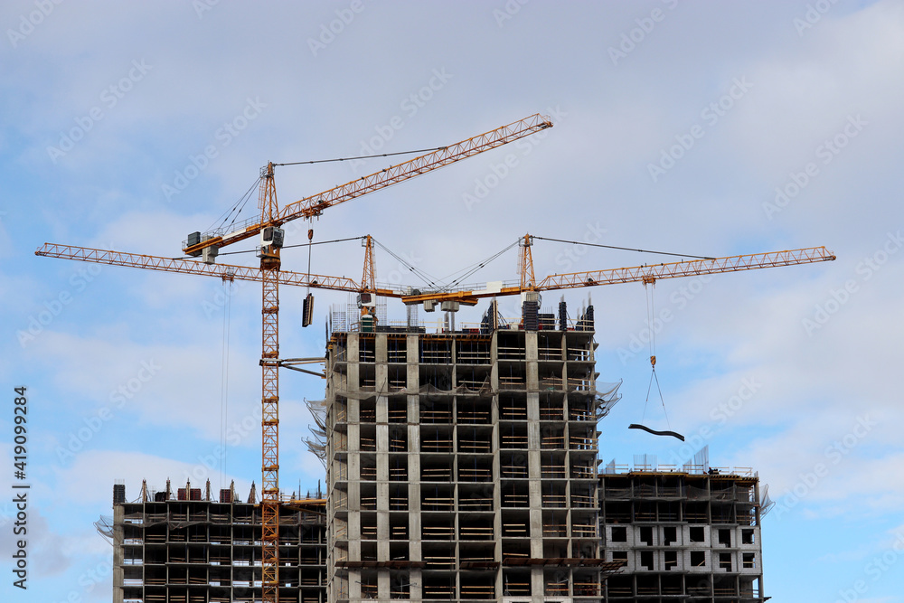 Construction cranes lifting the cargo above unfinished residential buildings on blue sky and white clouds background. Housing construction, apartment block in city