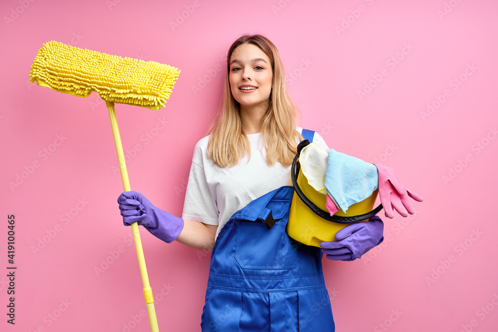 Smiling nice caucasian woman in blue coveralls and rubber gloves look at camera holding cleaning supplies. isolated pink background, portrait of pretty female ready to clean Smiling nice caucasian woman in blue coveralls and rubber gloves look at camera holding cleaning supplies. isolated pink background, portrait of pretty female ready to clean