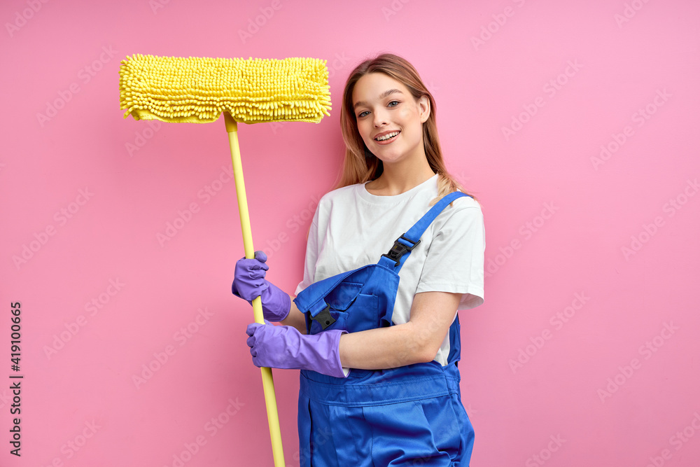 pleasant house wife in good mood holding cleaning equipment, rag for floor, wearing blue uniform coveralls isolated over pink studio background. portrait pleasant house wife in good mood holding cleaning equipment, rag for floor, wearing blue uniform coveralls isolated over pink studio background. portrait