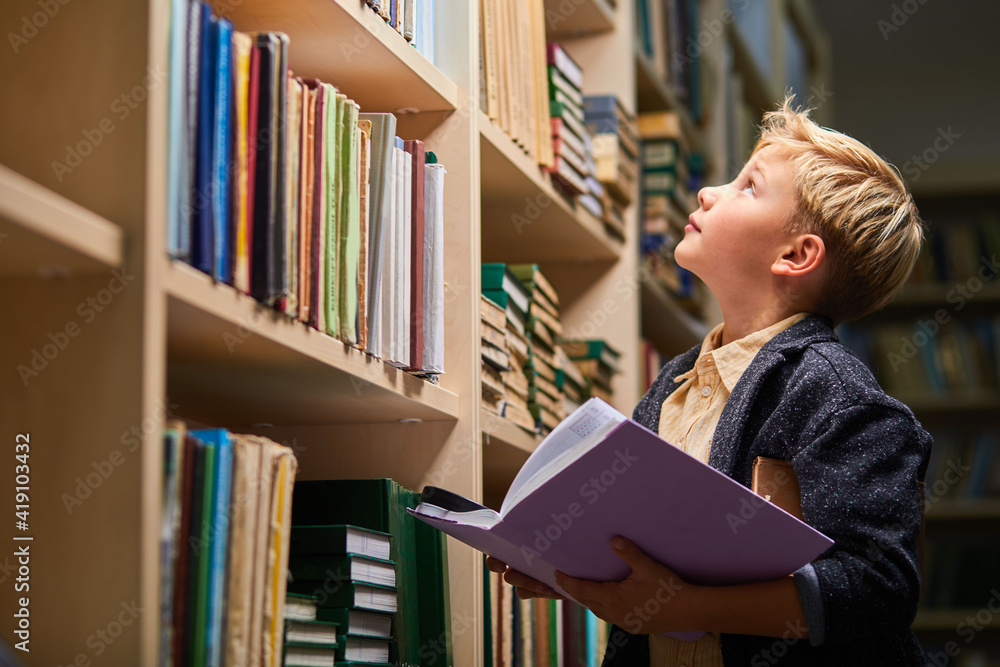 school boy looking up at bookshelves in campus library, child boy holding book in hands school boy looking up at bookshelves in campus library, child boy holding book in hands