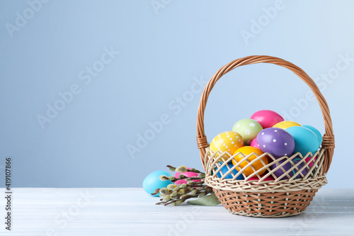 Colorful Easter eggs in wicker basket and willow branches on white wooden table. Space for text
