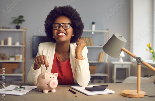 Money saving for shopping concept. Lovely satisfied, excited happy young african american woman in eyewear with yes gesture putting coin into piggybank because of income exceeds expenses significantly photo