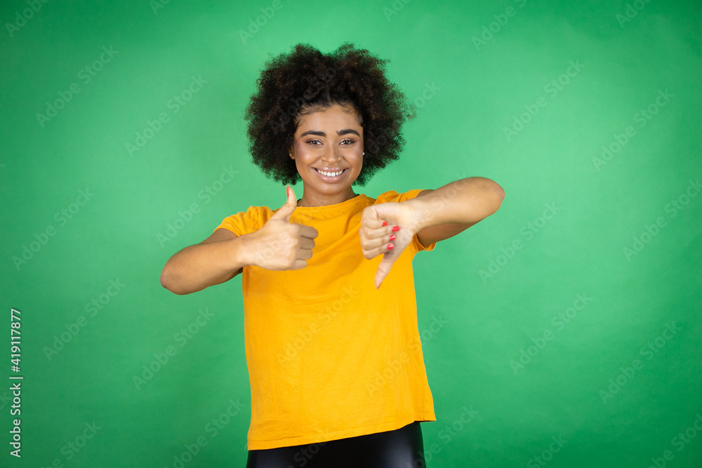 African american woman wearing orange casual shirt over green background Doing thumbs up and down, disagreement and agreement expression. Crazy conflict African american woman wearing orange casual shirt over green background Doing thumbs up and down, disagreement and agreement expression. Crazy conflict