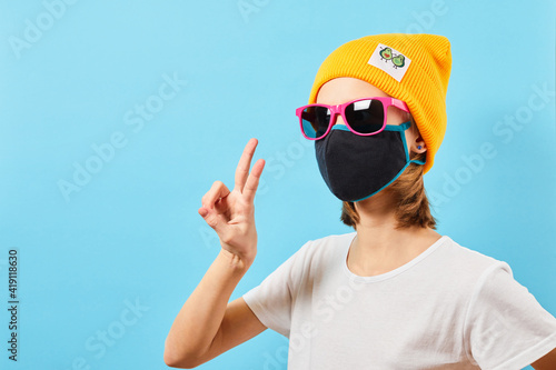 Hipster Teen in trendy glasses wearing knitted hat showing peace gesture. Young girl on blue background. Teenager Looking camera and showing v-sign symbols.