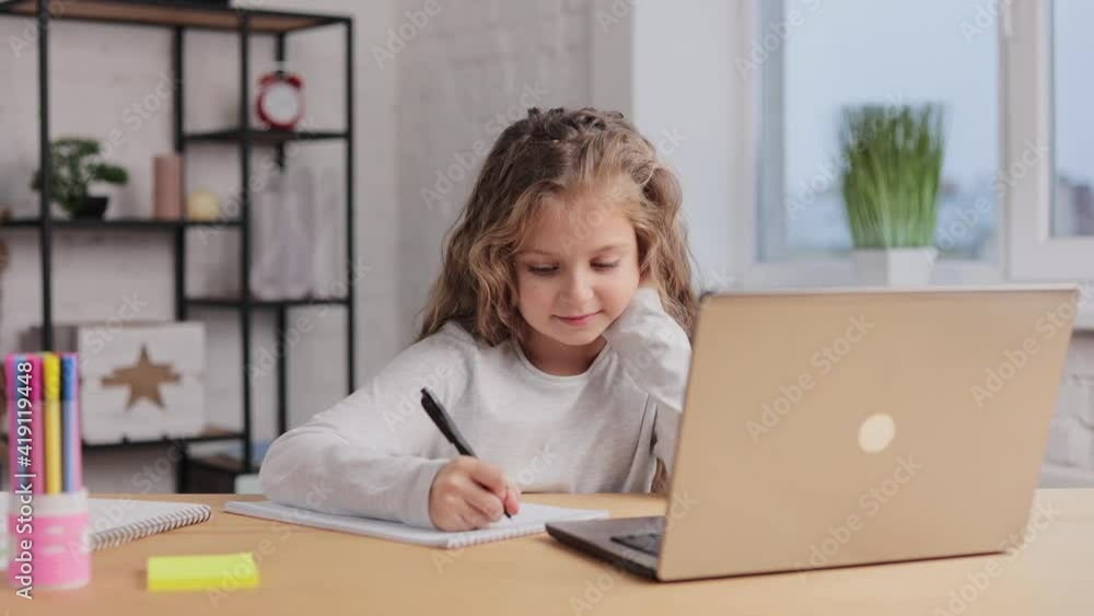 Cute primary school girl studying at home using laptop computer. Schoolgirl has online lesson, writing in exercise book, doing homework. Distance learning online. Children home education Cute primary school girl studying at home using laptop computer. Schoolgirl has online lesson, writing in exercise book, doing homework. Distance learning online. Children home education