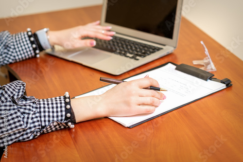 Female hands holding pen under the document and working with laptop at the table in an office