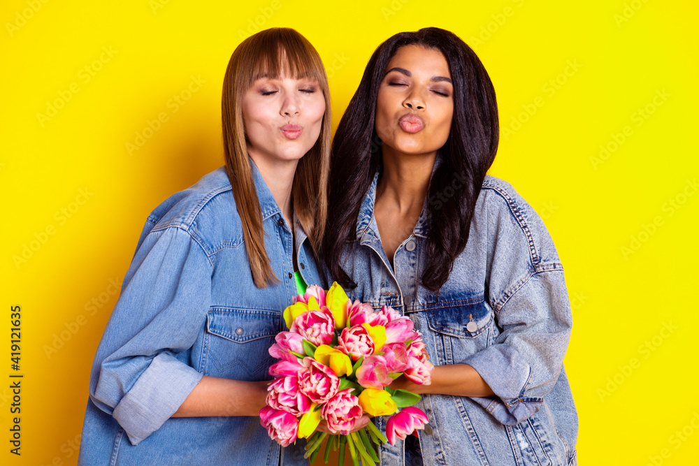 Photo of adorable ladies closed eyes kiss lips arms hold fresh flowers isolated on yellow color background Photo of adorable ladies closed eyes kiss lips arms hold fresh flowers isolated on yellow color background