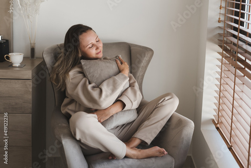 Caucasian woman dressed in comfy grey loungewear sitting in a cozy armchair in her bedroom and holding a decorative pillow. Coziness and comfort concept