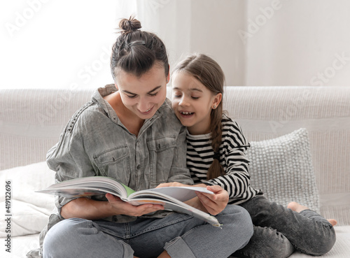 Young mother and daughter have fun together, reading a book at home on the couch.