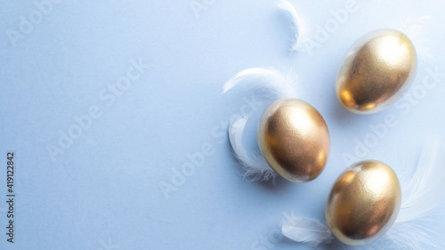 Golden easter colour eggs with white feathers on pastel blue background in Happy Easter decoration. Spring holiday top view concept.
