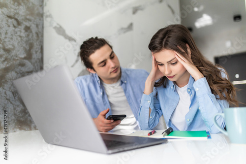 Young couple with many debts doing paperwork together, reviewing their bills, planning family budget and calculating finances at kitchen table with papers, calculator and laptop