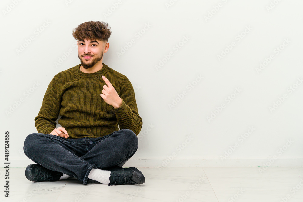 Young Moroccan man sitting on the floor isolated on white background pointing with finger at you as if inviting come closer.