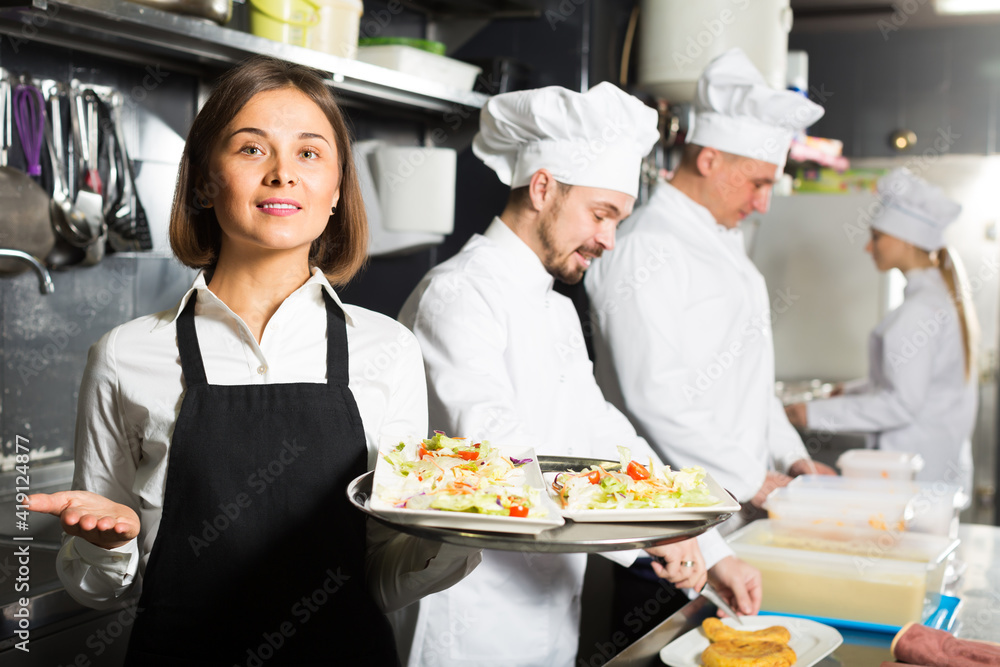 Laughing female waiter taking ordered dishes from restaurant kitchen