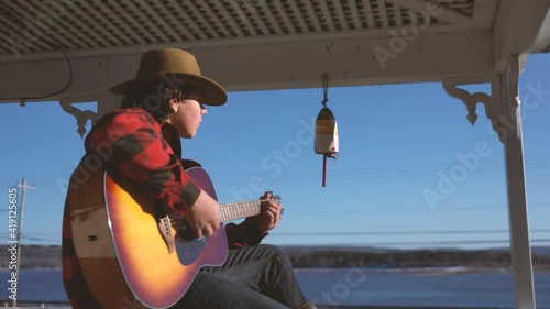 A teenager playing acoustic guitar outside on a deck overlooking the sea.
