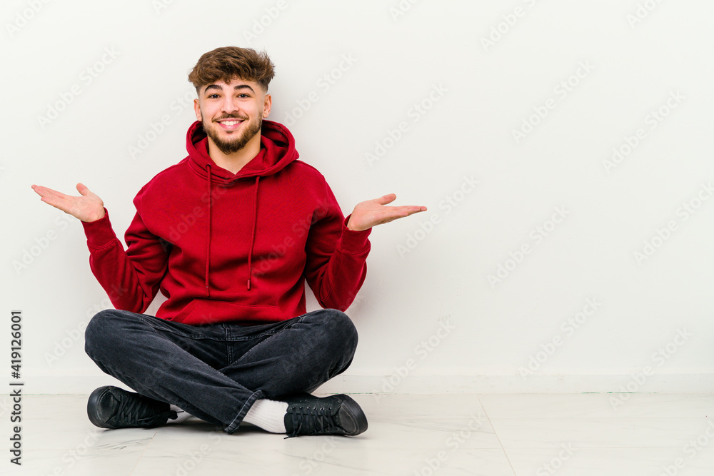 Young Moroccan man sitting on the floor isolated on white background makes scale with arms, feels happy and confident. Young Moroccan man sitting on the floor isolated on white background makes scale with arms, feels happy and confident.
