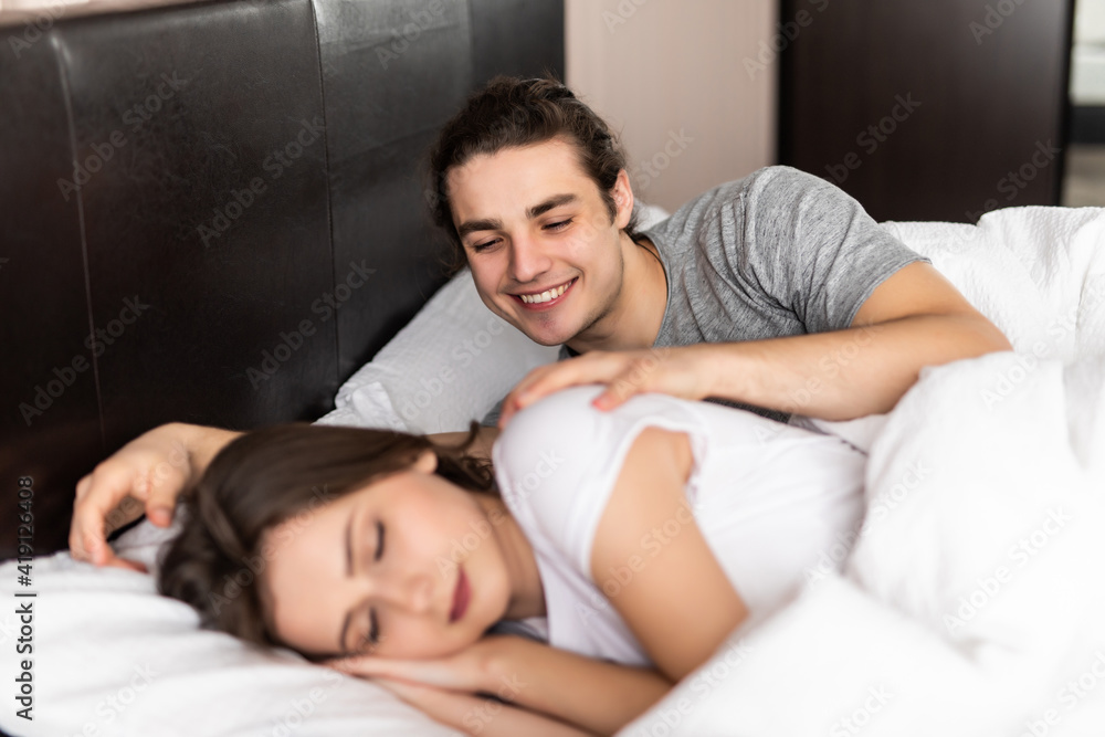 Happy handsome young man lying and waking his woman in bed. Couple wake up together. Happy handsome young man lying and waking his woman in bed. Couple wake up together.