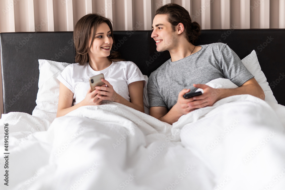 Portrait of a jelous young man looking at his girlfriends mobile phone while sitting together in bed at home Portrait of a jelous young man looking at his girlfriends mobile phone while sitting together in bed at home