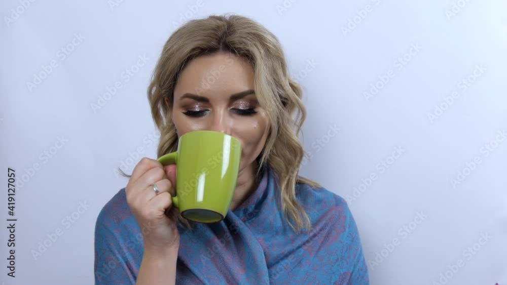 Beautiful young woman model holds green cup of tea or coffee in hands, drinks it enjoys and looks pensively on white background. Young girl with beautiful smoky eye makeup and blue scarf on shoulders. Beautiful young woman model holds green cup of tea or coffee in hands, drinks it enjoys and looks pensively on white background. Young girl with beautiful smoky eye makeup and blue scarf on shoulders.