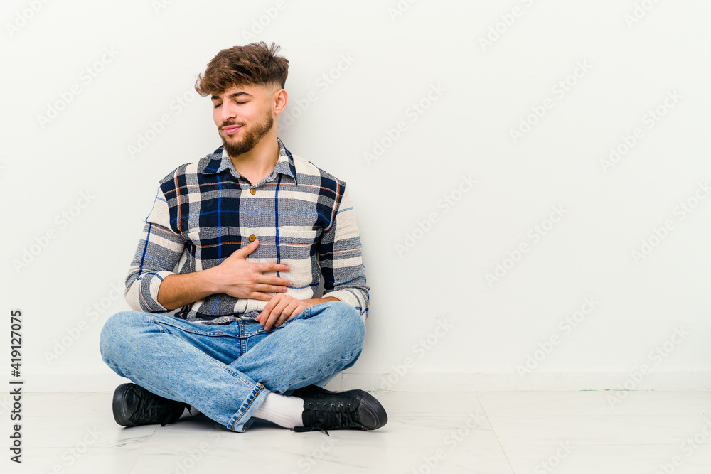 Young Moroccan man sitting on the floor isolated on white background touches tummy, smiles gently, eating and satisfaction concept.