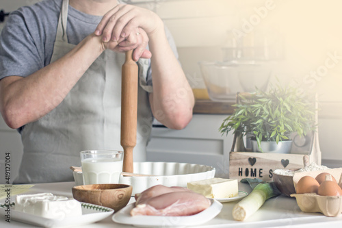 A male cook in an apron holds a rolling pin for rolling out dough in front of the kitchen table with ingredients for the pie. Home cooking an authentic hobby