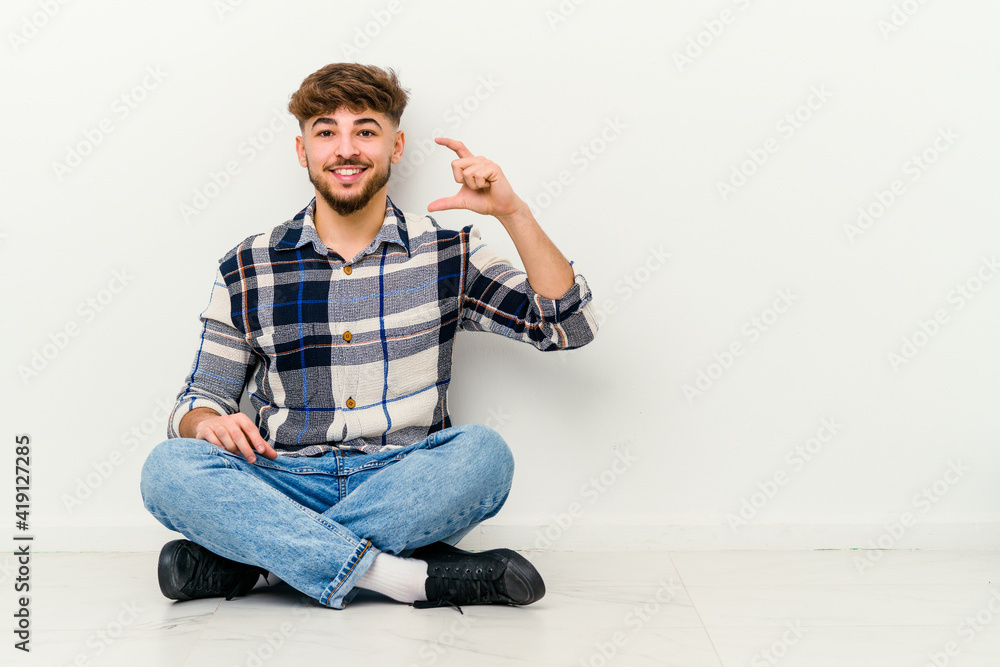 Young Moroccan man sitting on the floor isolated on white background holding something little with forefingers, smiling and confident. Young Moroccan man sitting on the floor isolated on white background holding something little with forefingers, smiling and confident.