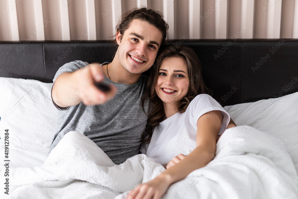 People, rest, love, relationships and television concept. Happy couple with remote lying in bed at home and watching tv People, rest, love, relationships and television concept. Happy couple with remote lying in bed at home and watching tv