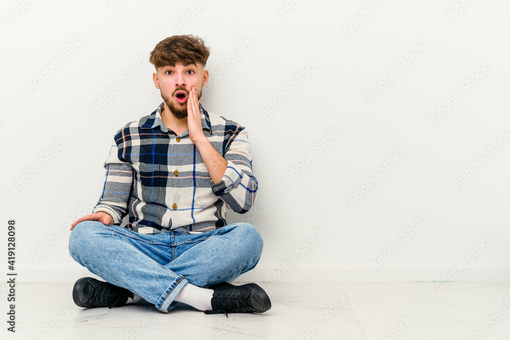Young Moroccan man sitting on the floor isolated on white background shouts loud, keeps eyes opened and hands tense.