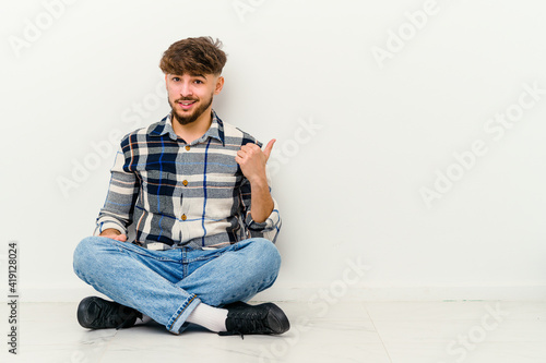 Young Moroccan man sitting on the floor isolated on white background shocked pointing with index fingers to a copy space.