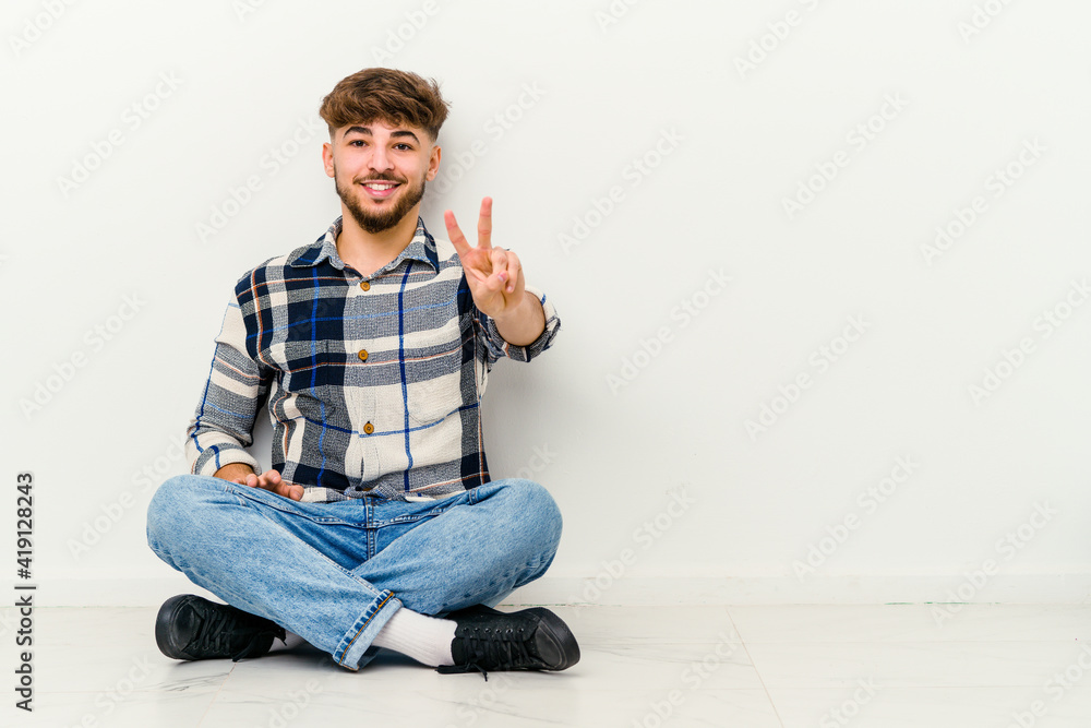 Young Moroccan man sitting on the floor isolated on white background showing victory sign and smiling broadly.