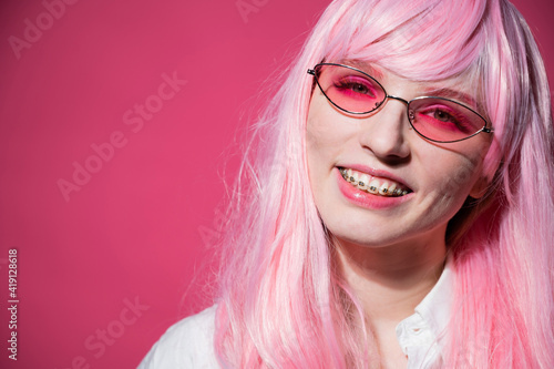 Close-up portrait of a young woman with braces in a pink wig and sunglasses on a pink background.