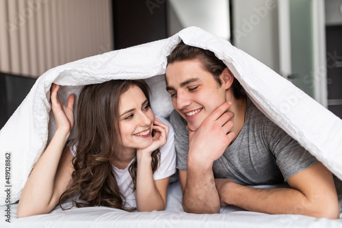 Cheerful young couple head against head under the duvet in bed