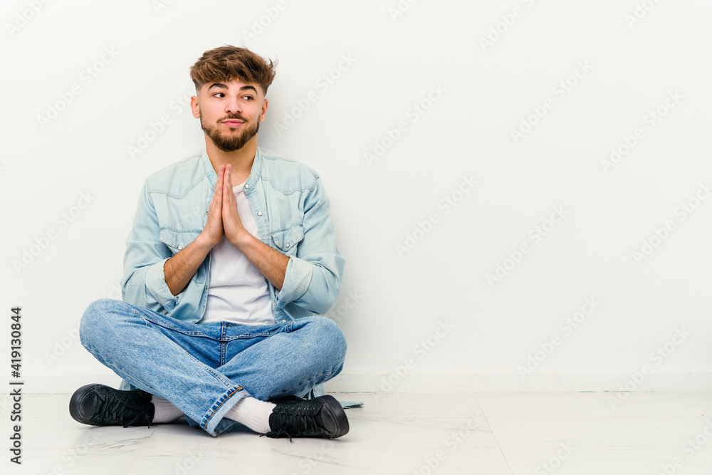 Young Moroccan man sitting on the floor isolated on white background praying, showing devotion, religious person looking for divine inspiration. Young Moroccan man sitting on the floor isolated on white background praying, showing devotion, religious person looking for divine inspiration.
