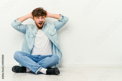 Young Moroccan man sitting on the floor isolated on white background screaming, very excited, passionate, satisfied with something. © Asier