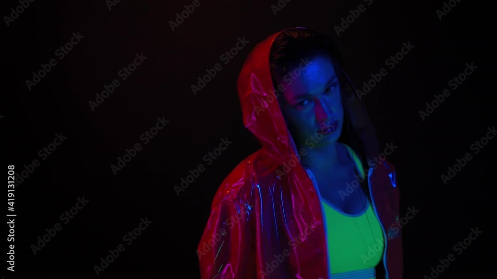Turning on camera in a glossy jacket hood slow motion girl moving in neon light on black background. Beautiful girl dancing in lights wearing sunglasses. Night club, Party. FHD footage. Turning on camera in a glossy jacket hood slow motion girl moving in neon light on black background. Beautiful girl dancing in lights wearing sunglasses. Night club, Party. FHD footage.