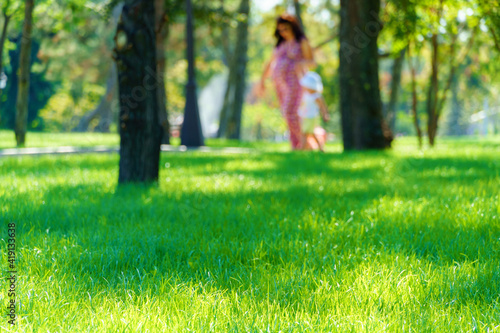 woman and child walk in a city park, summer day, green lawn with grass and trees, bright sunlight and shadows