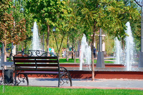 city park on a summer day, green lawns with grass and trees, path, benches and fountain, bright sunlight and shadows