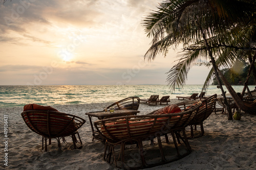 Fototapeta Naklejka Na Ścianę i Meble -  wood chairs and hammock on a background of a beautiful small island and wight beaches on big ocean with trees and palms of coconut and a part of a local port and bungalow wood before the sunset