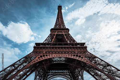 Ultra wide angle of Eiffel Tower over blue sky with white clouds and bright glow of sun in Paris, France. Worms eye view of the entire monument with all little details. Scene with high dynamic range.