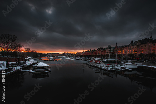 Red sunset with dark clouds over the pier of the winter city (Stockholm). The waterfront is lined with historic buildings and a fine snow cover is visible in the surroundings.