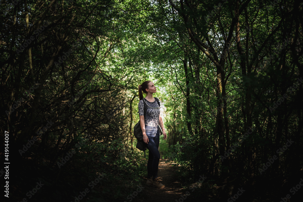 Young woman standing in middle of dense vegetation forming narrow alley. Soft light shines through vault of the branches and leaves. Woman is casually dressed with hydrating backpack on her back. Young woman standing in middle of dense vegetation forming narrow alley. Soft light shines through vault of the branches and leaves. Woman is casually dressed with hydrating backpack on her back.