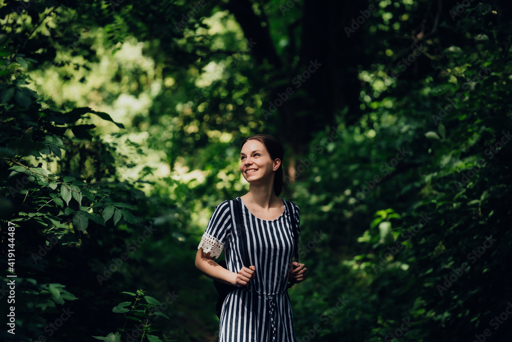 Beautiful young girl with small backpack and positive expression on her face. She is dressed in dress with striped pattern and white lace. It is located in middle of forest with subtle tones of light. Beautiful young girl with small backpack and positive expression on her face. She is dressed in dress with striped pattern and white lace. It is located in middle of forest with subtle tones of light.