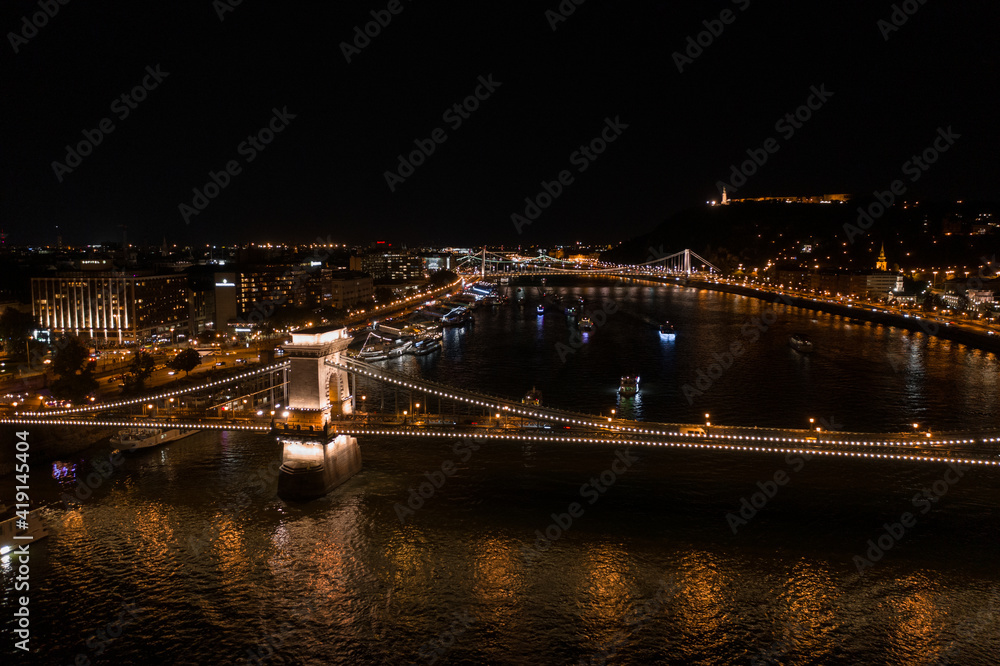 Aerial view of illuminated Budapest Chain Bridge at night with dark sky and reflection in Danube river. Panoramic view of hungarian famous bridge. Budapest, Hungary at night.