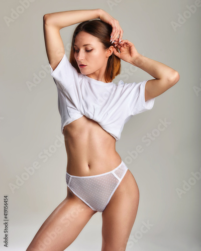 Beautiful, seductive woman posing in sexy white underpants and t-shirt. Sporty figure on gray background. Seductive woman proud of her perfect body. Sexy white erotic underwear.
