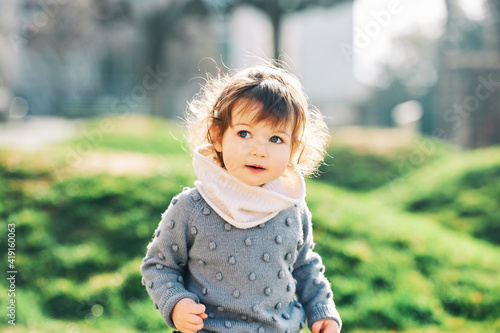 Outdoor portrait of adorable 1 - 2 year old toddler girl, wearing knitted pullover, playing in city park, early spring weather