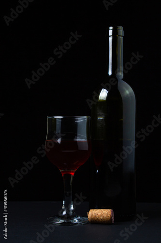 A glass of homemade wine next to a bottle on a dark background. Glass of red wine on a black background. Alcoholic drink