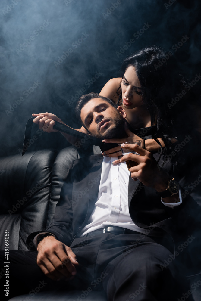Man in suit holding whiskey near sexy woman on couch on black background with smoke