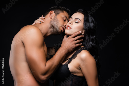 Shirtless man touching neck and kissing sexy woman isolated on black